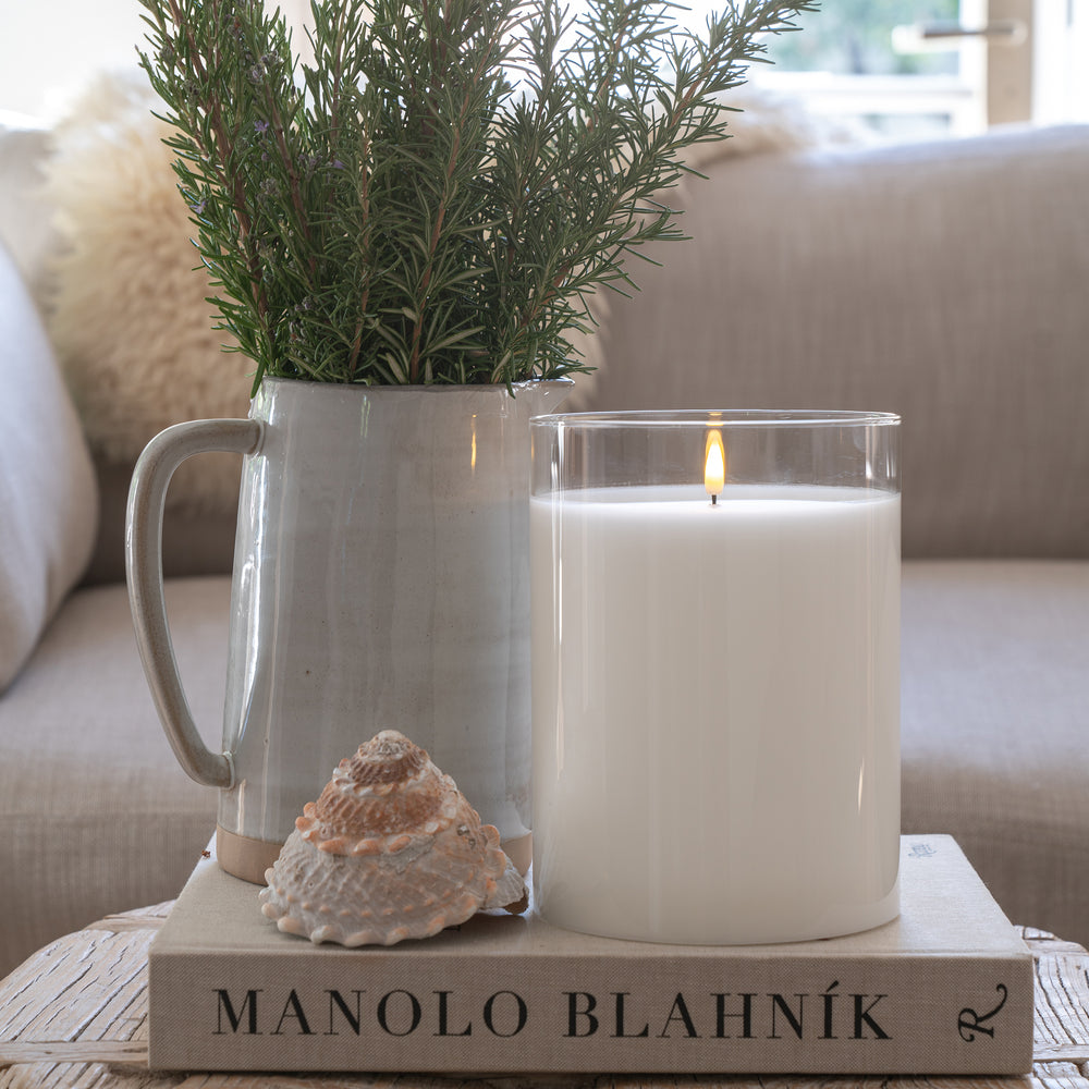 Elegant white flameless LED hurricane candle glows softly on a book beside a delicate seashell, with a fresh rosemary bouquet in a tall vase. A Restoration Hardware couch adds a cozy, luxurious backdrop, creating a serene and sophisticated home ambiance.