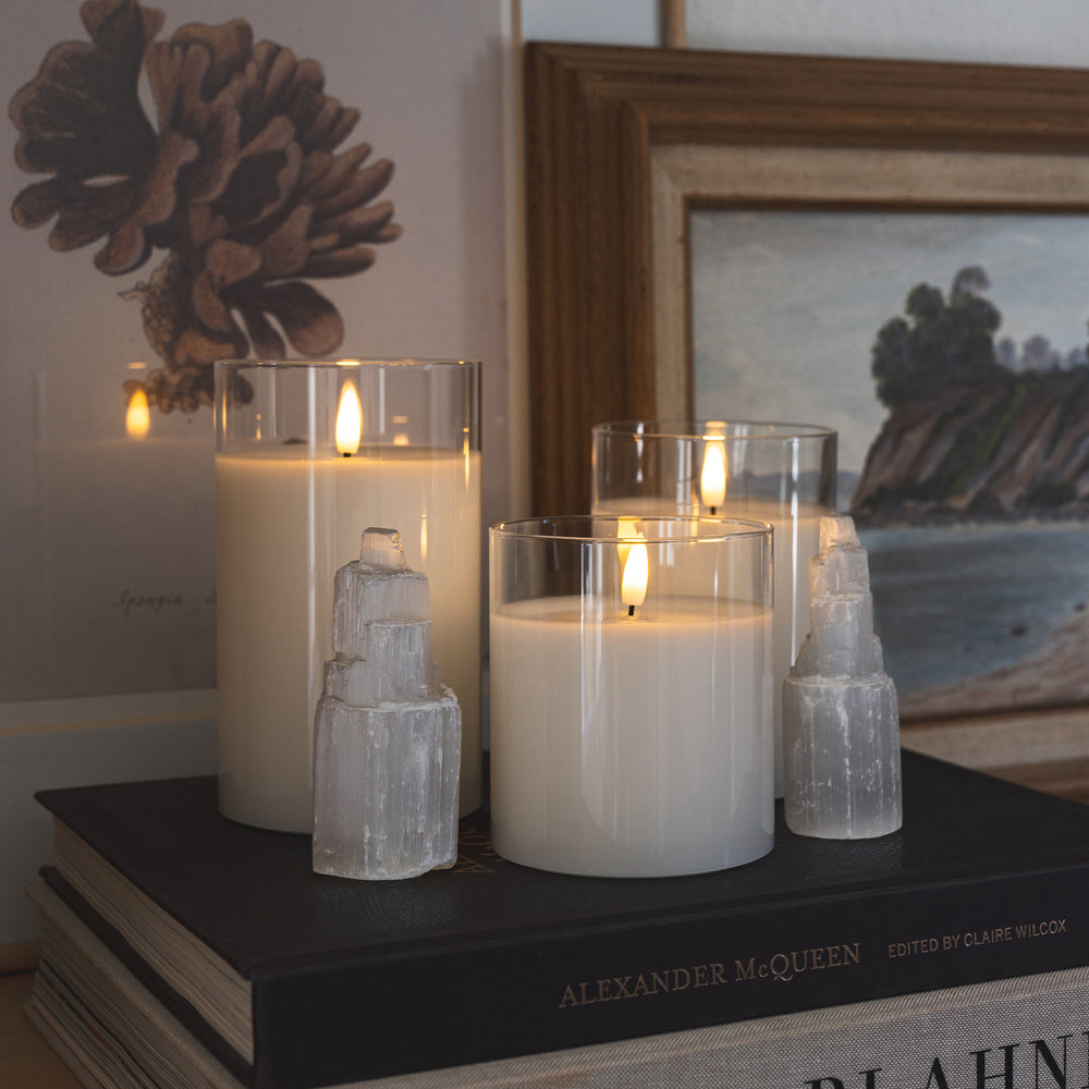 Three hurricane glass flameless white candles sit atop two stacked books on a shelf. A piece of artwork hangs in the background, adding a stylish touch. The flameless candles provide a warm glow and are safe for children and pets.